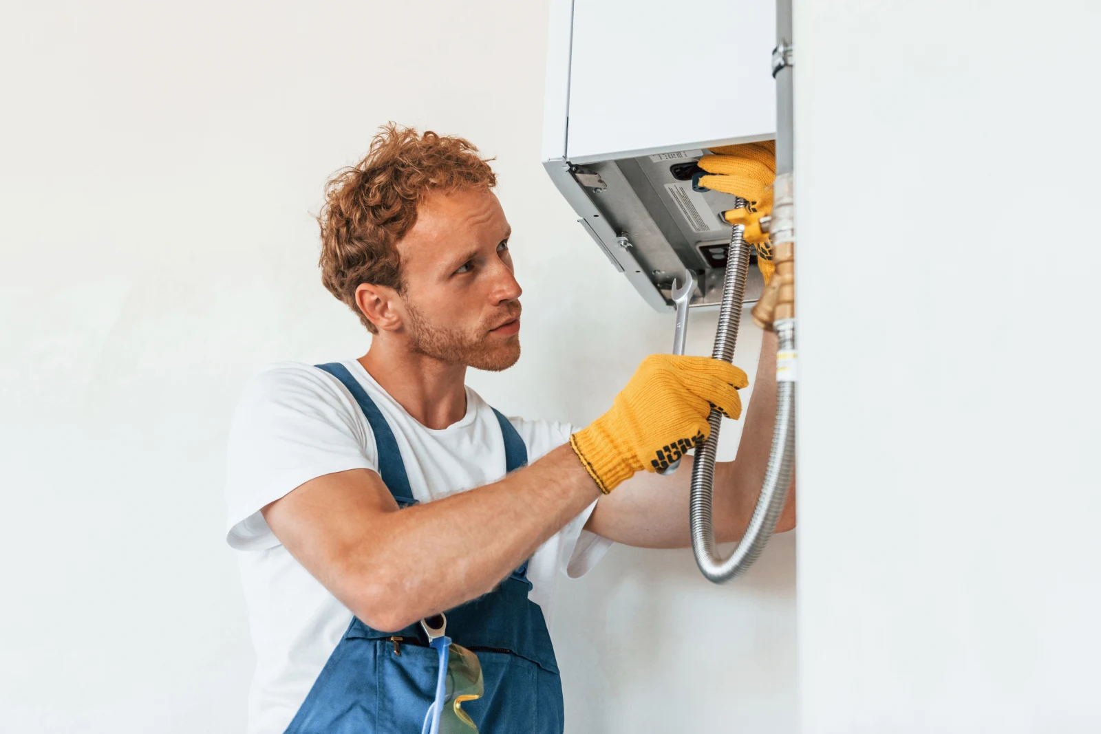 A boiler repair technician in work gloves using a wrench on a boiler's pressure fittings and hose during a descale service.