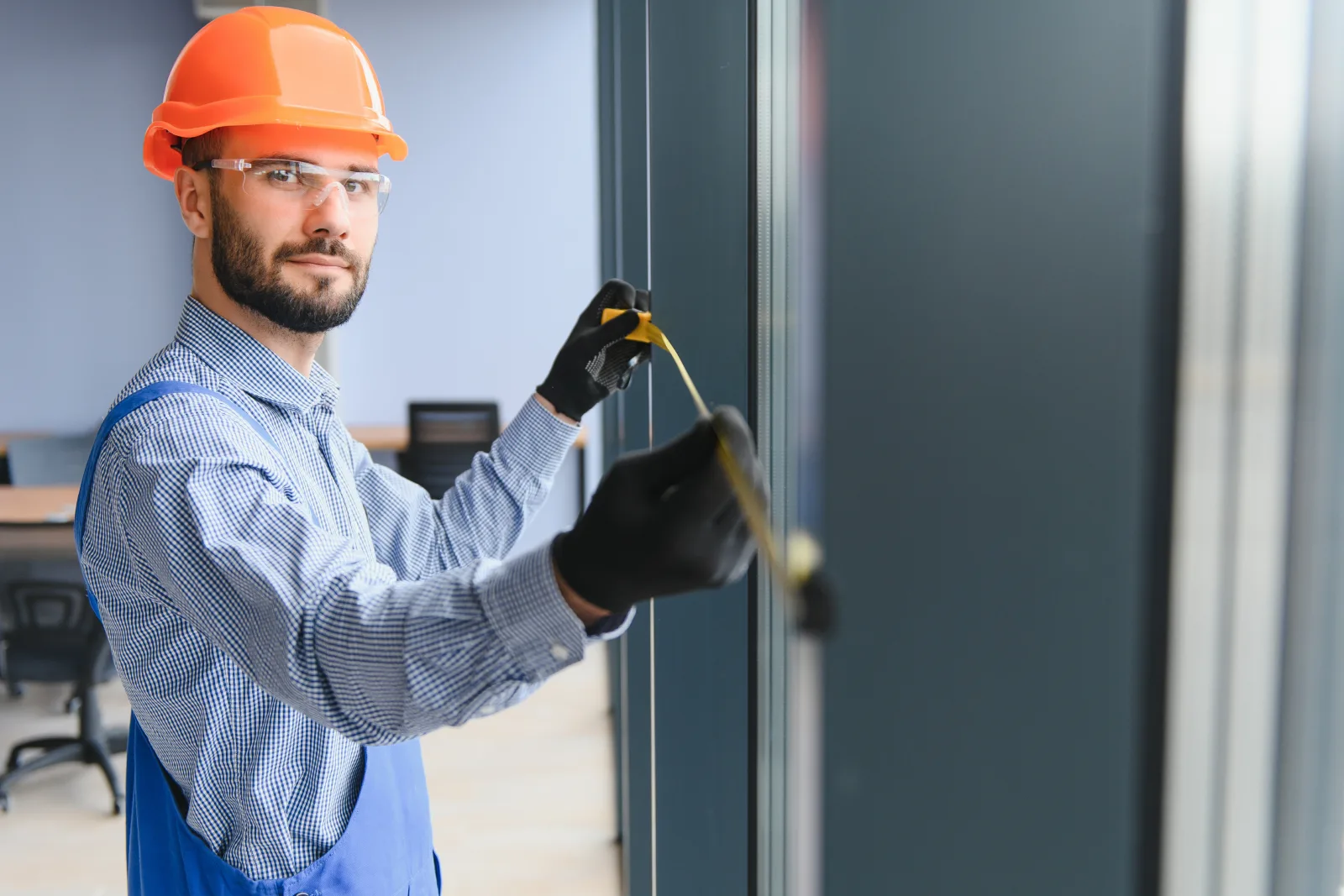A technician in a hard hat and safety glasses measuring a glass storefront door frame ahead of a commercial door hardware installation in a Phoenix office building.