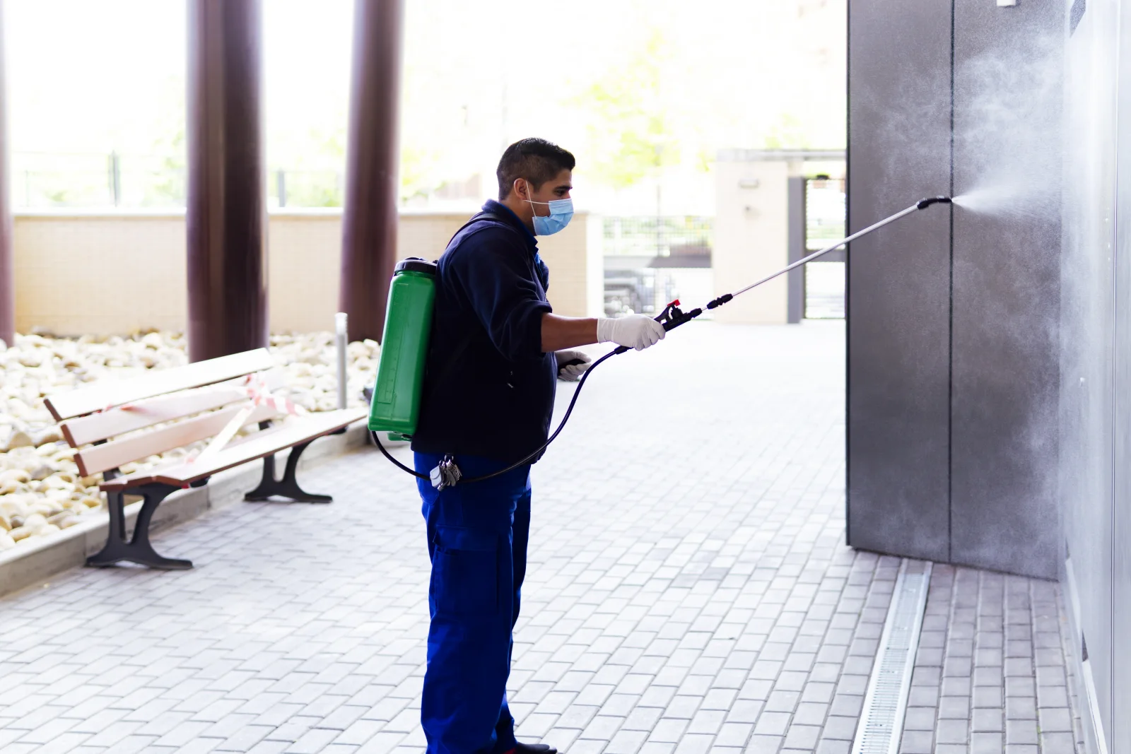 A pest control technician in uniform treating the exterior wall of a Phoenix commercial building with a backpack sprayer.