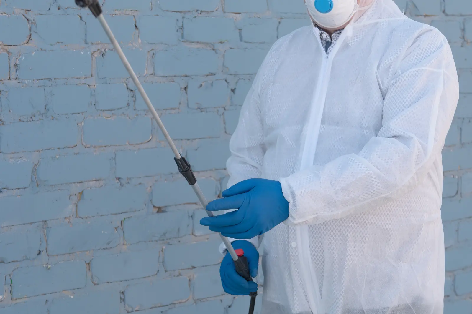 A licensed pest control technician in full protective gear applying a low-toxicity treatment to a home's exterior wall.