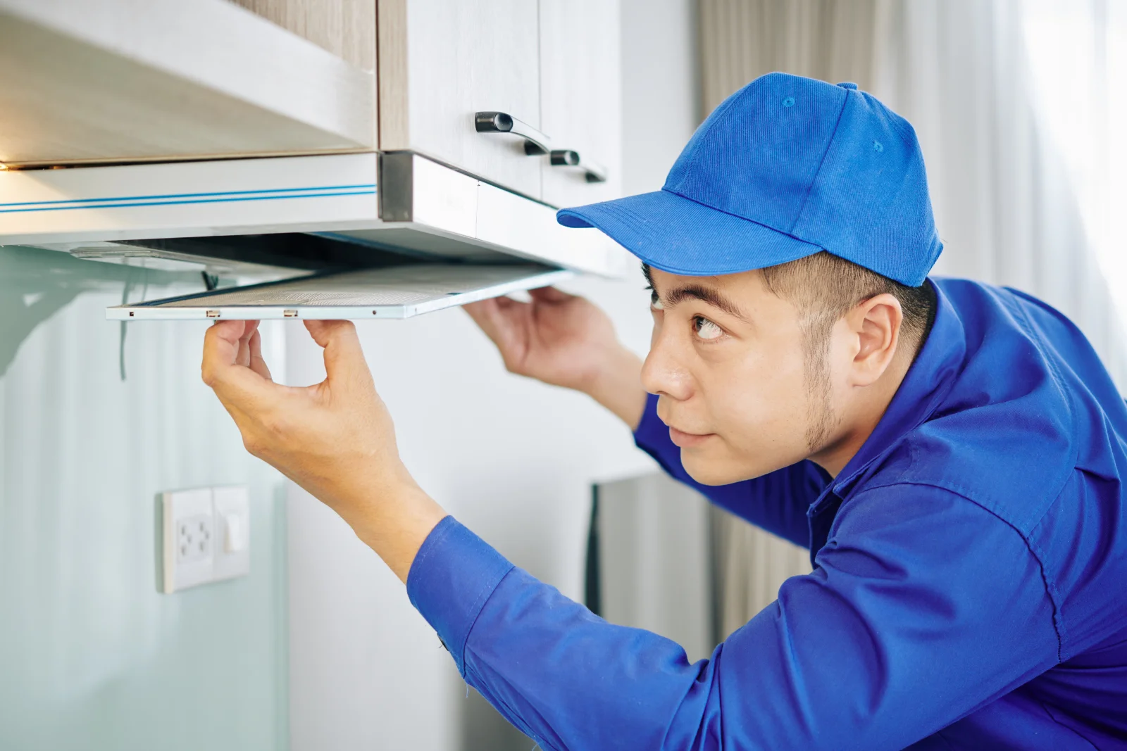 A technician in a blue uniform inspecting a kitchen range hood exhaust fan and filter, the kind of diagnosis a Phoenix appliance pro runs before quoting a fan repair.