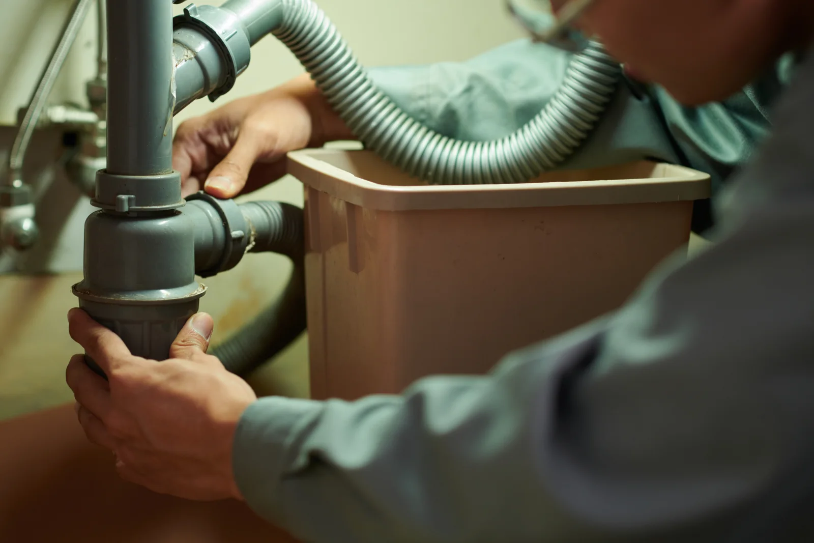 grease-trap-cleaning-phoenix-technician-connecting-drain-line Grease trap cleaning in Phoenix, Arizona — a plumber's gloved hands connecting a drain line and pump hose beneath a commercial kitchen sink.