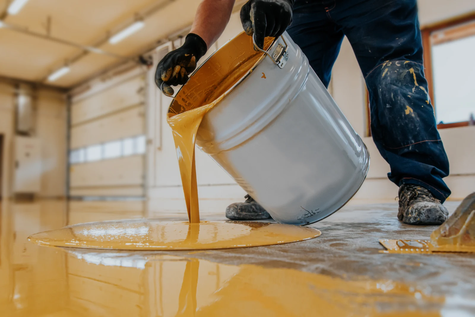 industrial-paint-contractors-phoenix-floor-coating A worker pouring fresh epoxy floor coating onto a concrete warehouse floor in Phoenix, Arizona before spreading it into a seamless protective surface.