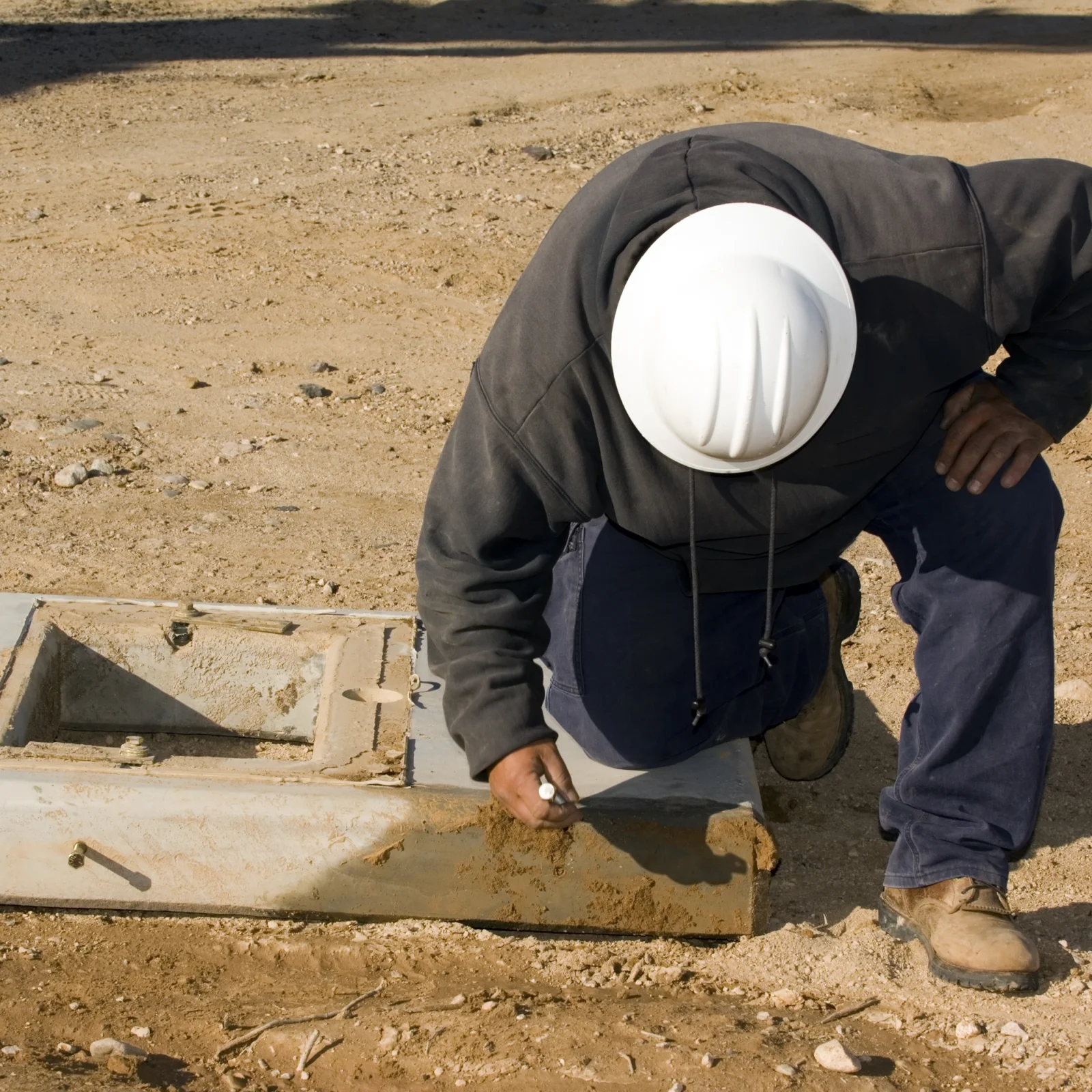 landscape-drainage-phoenix-catch-basin-inspection Landscape drainage in Phoenix, Arizona — a technician in a hard hat inspecting a concrete drainage catch basin set into bare desert soil.