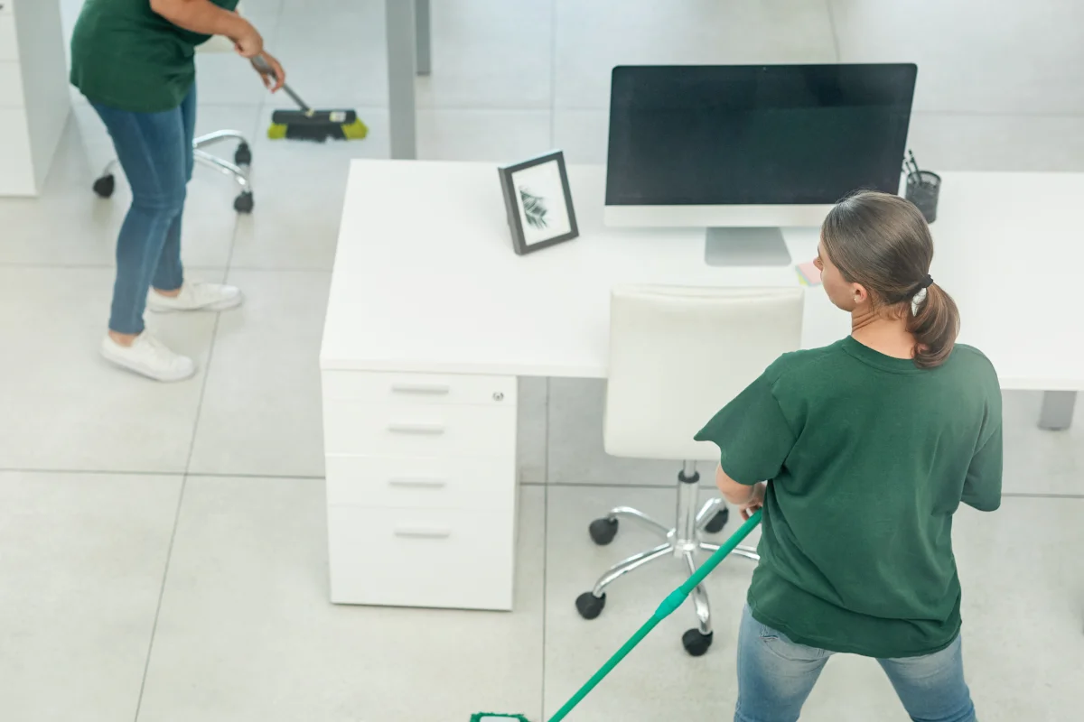 office-cleaning-services-phoenix-az-team-mopping-modern-office Office cleaning services in Phoenix, AZ — an aerial view of two cleaners in green branded uniforms mopping floors and wiping desks in a modern open-plan office.