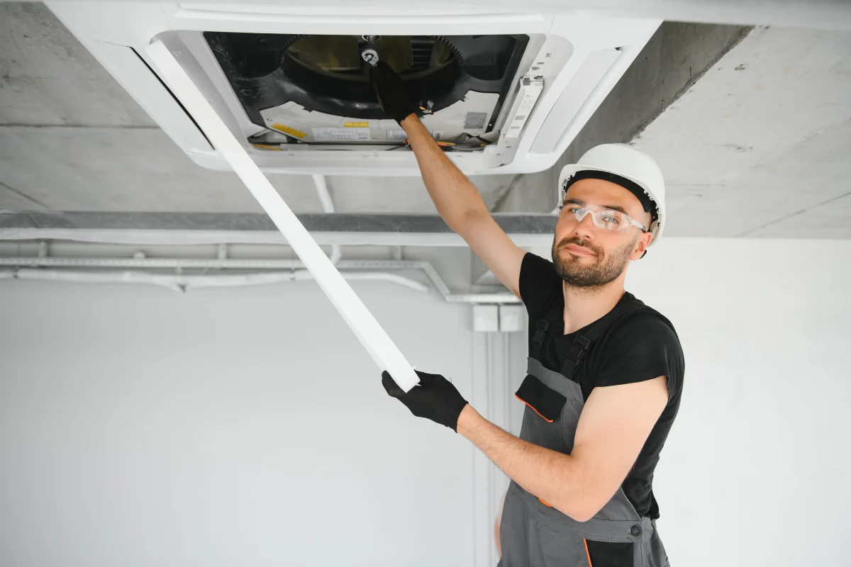 phoenix-duct-cleaning-vent-access Phoenix duct cleaning — a uniformed HVAC technician in a hard hat and safety glasses accessing a ceiling duct vent to inspect and clean the ductwork.