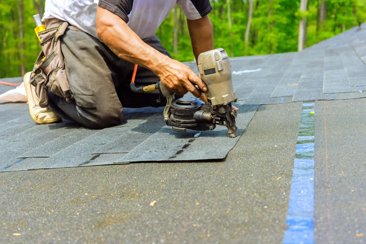 reroofing-phoenix-shingle-installation-nail-gun Reroofing in Phoenix, Arizona — a professional roofer in a tool belt kneeling on a roof and using a nail gun to install new shingles.