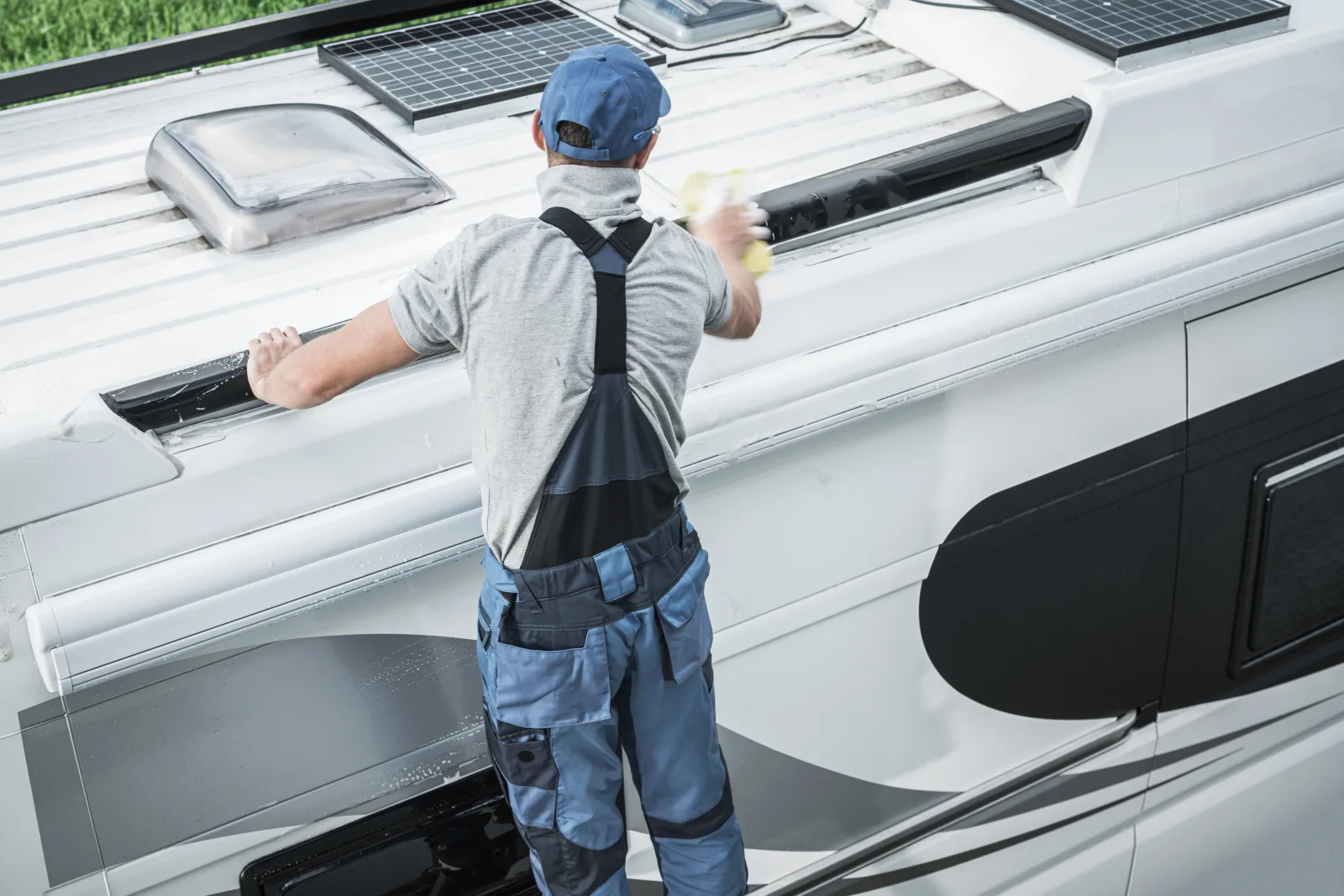 A technician on an RV roof resealing a seam near a solar panel and roof vent.