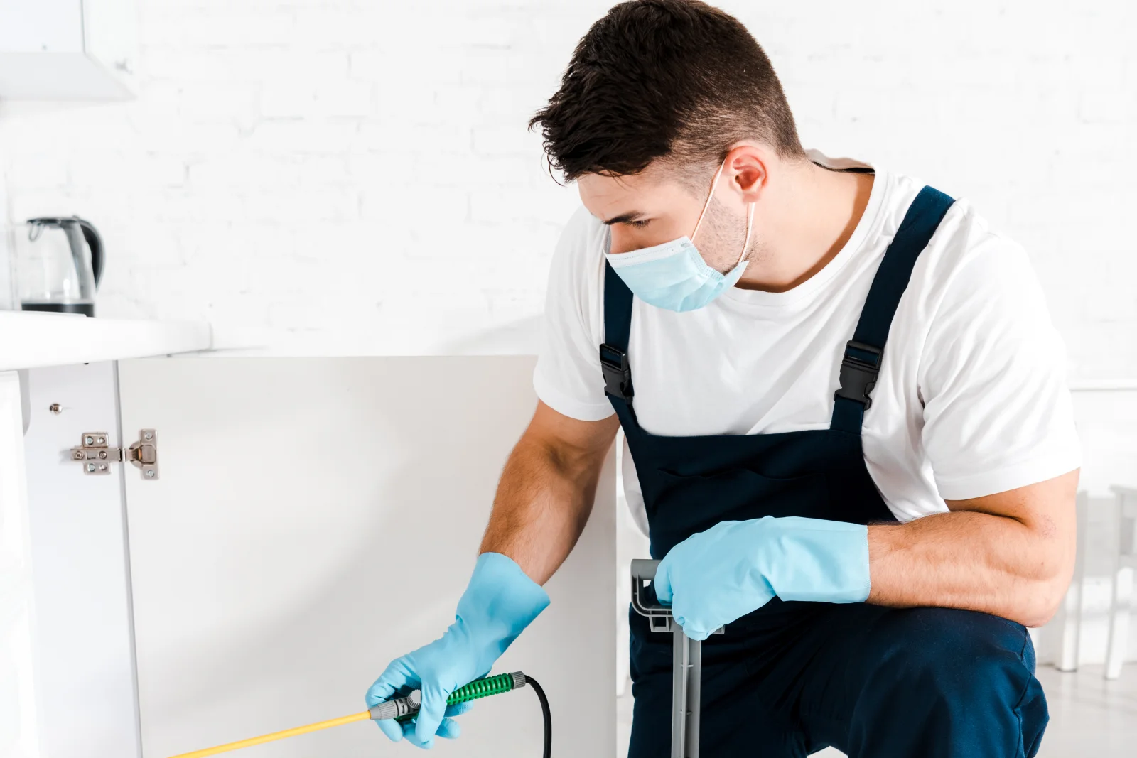 Silverfish control in Phoenix, Arizona — a pest control technician in a mask and gloves treating under a kitchen cabinet near a moisture source.
