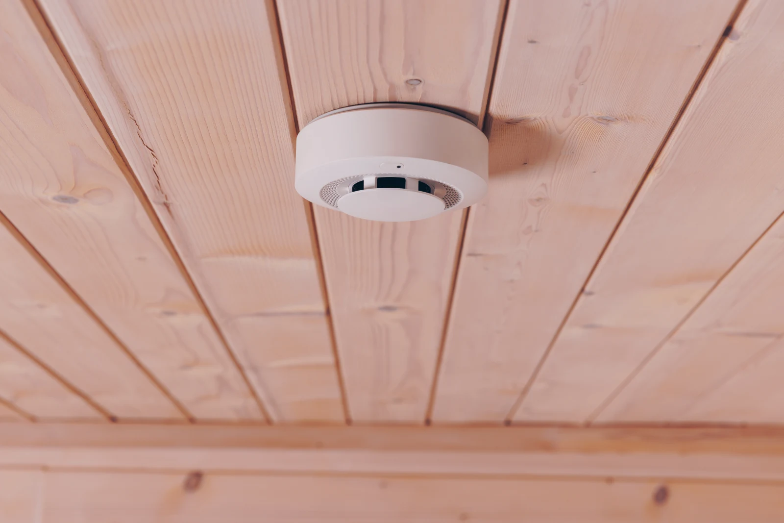 smoke-detector-repair-phoenix-attic-heat A smoke detector mounted on a wood ceiling beneath an attic space, where Phoenix heat and dust cause false alarms and failures.