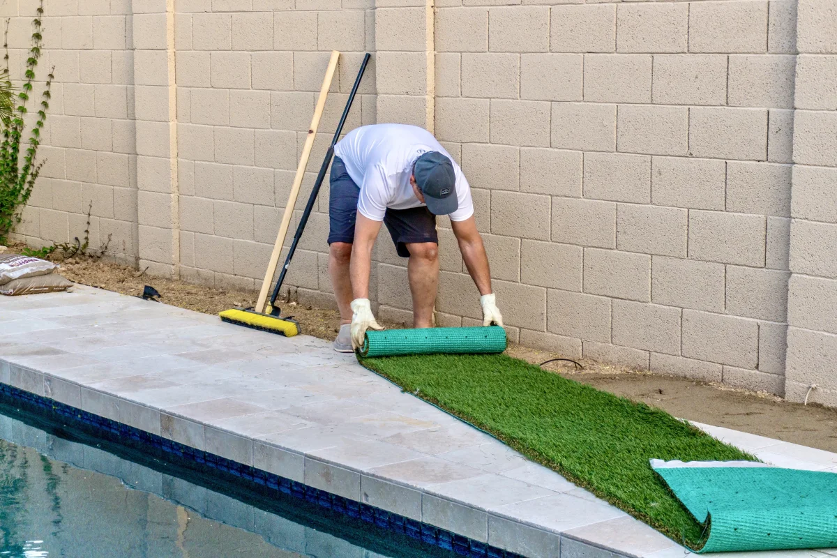 turf-installation-phoenix-poolside-artificial-grass Turf installation in Phoenix, Arizona — a professional installer in a polo, cap, and gloves rolling out artificial turf along a poolside block wall.