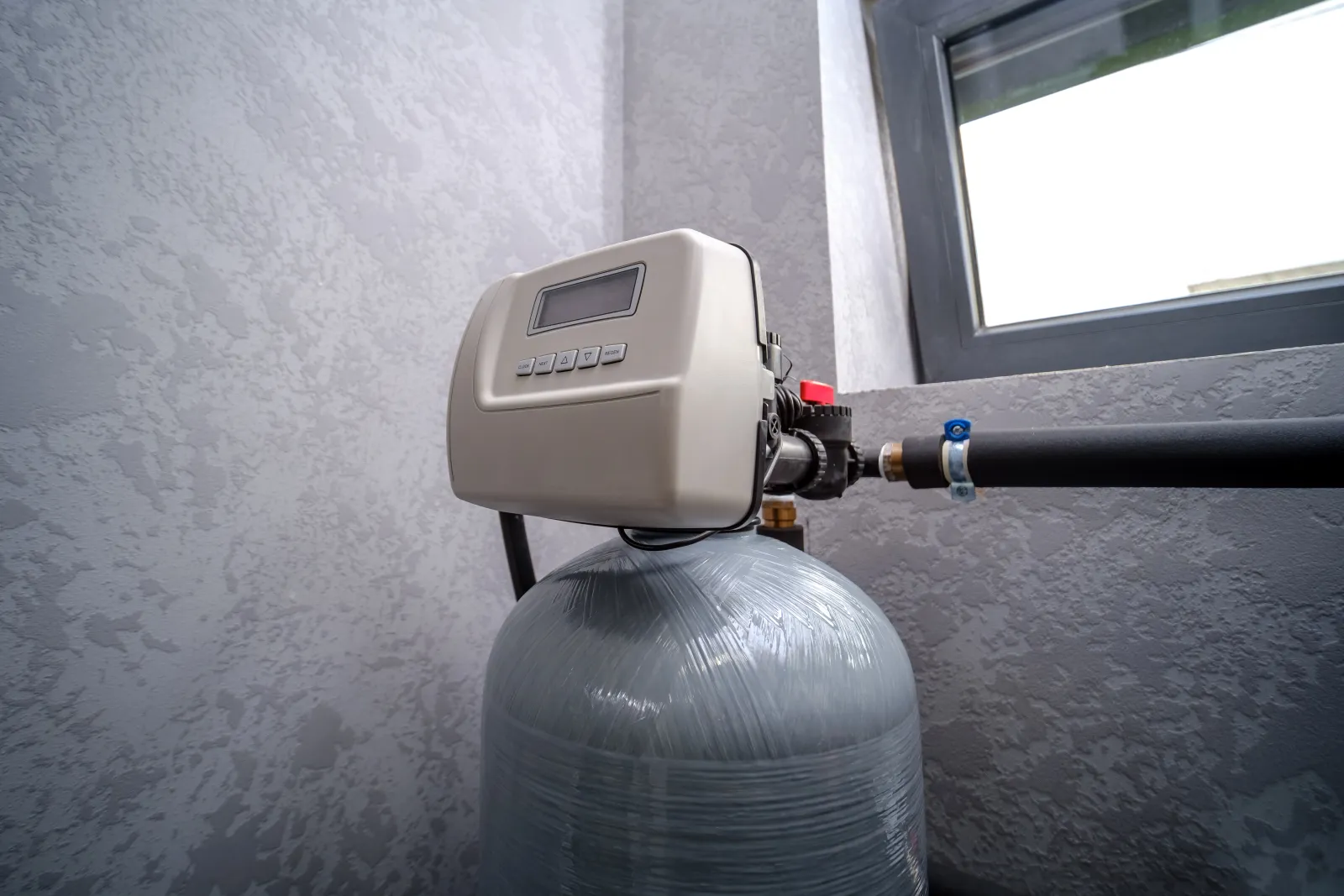A residential water softener with its digital control valve mounted on the resin tank beside a Phoenix home window, the kind of unit a water-treatment pro services when soft water stops flowing.